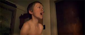 yeenage boy with short hair and no shirt is shouting off-camera in a dimly lit room