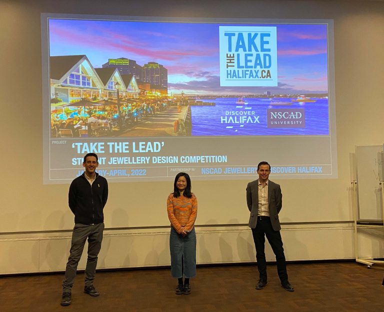 NSCAD faculty Greg Sims, design competition winner Meichan Yuan, and Discover Halifax representative Jeff Nearing standing in front of a PowerPoint backdrop with an image of Halifax and a text box that reads Take The Lead