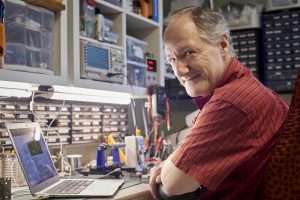 Michael LeBlanc sitting at his desk surrounded by tech devices, smiling at the camera