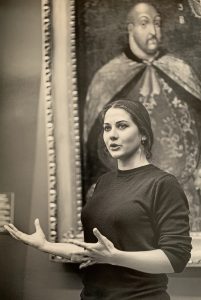 Woman standing in front of a painting with open hand gestures.
