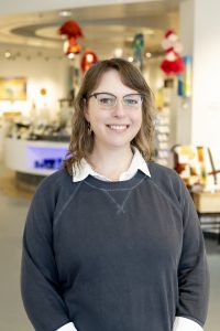 A woman wearing a blue sweatshirt and glasses stands in a gallery space surrounded by hand crafted items. She is turned slightly towards the camera, and she is smiling at the photographer.