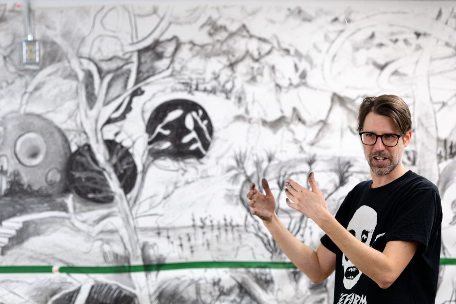 A man with dark hair and glasses stands in front of a black and white mural. He is wearing a black shirt with white drawing.