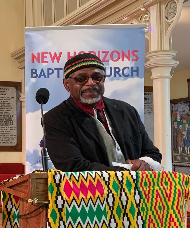 A Black man stands at a podium in front of a church sign. He is wearing a black jacket, shirt and tie, with glasses and African kufi cap