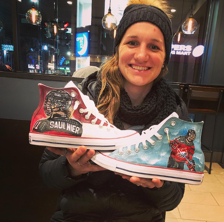 Canadian Olympic medalist, Jillian Saulnier, holds up sneakers customized by Smith. She is wearing a black beanie and outfit. The sneakers are painted blue and red with details from her hockey team uniform