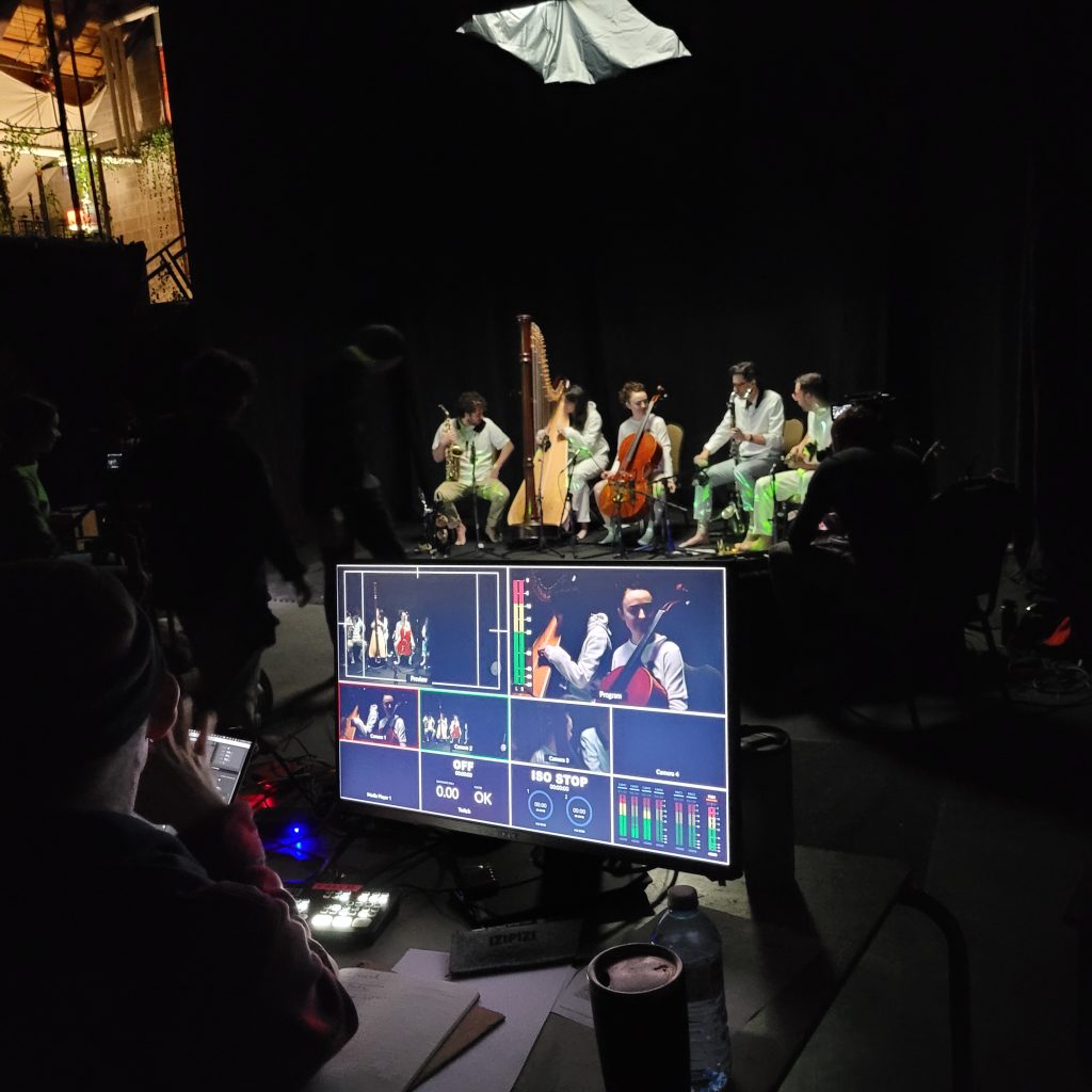 A group of technicians watch a screen monitor during the filming of a musical concert. The room is dark while the musicians are spotlighted with instruments in hand,