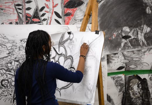 A black student with shoulder length hair draws on a canvas. She is wearing a black shirt and is using black charcoal on a white canvas