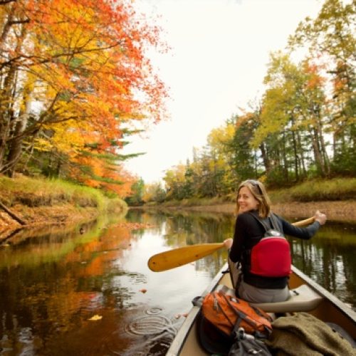 Person paddling a canoe on a river surrounded by autumn trees