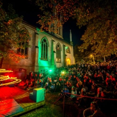 Crowd gathered at a night-time outdoor art event lit with coloured lights
